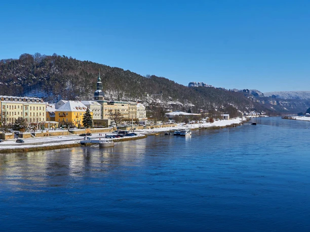 Blick auf das winterliche Bad Schandau Verschneite Stadtansicht am Flussufer mit bunten Gebäuden, einem Kirchturm und bewaldeten Hügeln im Hintergrund unter klarem, blauem Himmel.