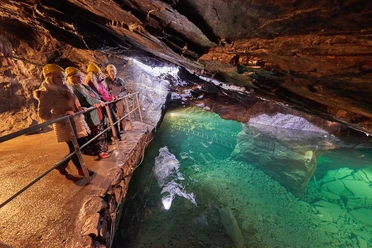 Führung im Besucherbergwerk "Marie Louise Stolln" Vier Personen mit gelben Helmen stehen auf einem Geländer in einer beleuchteten Höhle mit grünem Wasser, die Atmosphäre ist abenteuerlich.