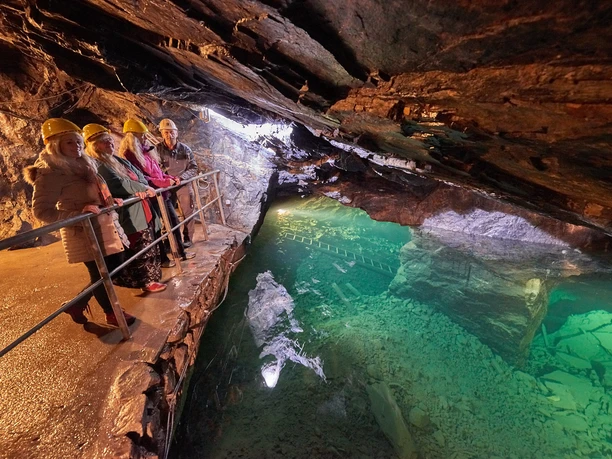 Führung im Besucherbergwerk "Marie Louise Stolln" Vier Personen mit gelben Helmen stehen auf einem Geländer in einer beleuchteten Höhle mit grünem Wasser, die Atmosphäre ist abenteuerlich.