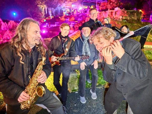 Farbenbaden Miniaturpark "Kleine Sächsische Schweiz" Eine Gruppe von Musikern spielt nachts im Freien; ein Mann mit Saxophon, ein anderer mit Gitarre, umgeben von bunten Lichtern und fröhlicher Stimmung.