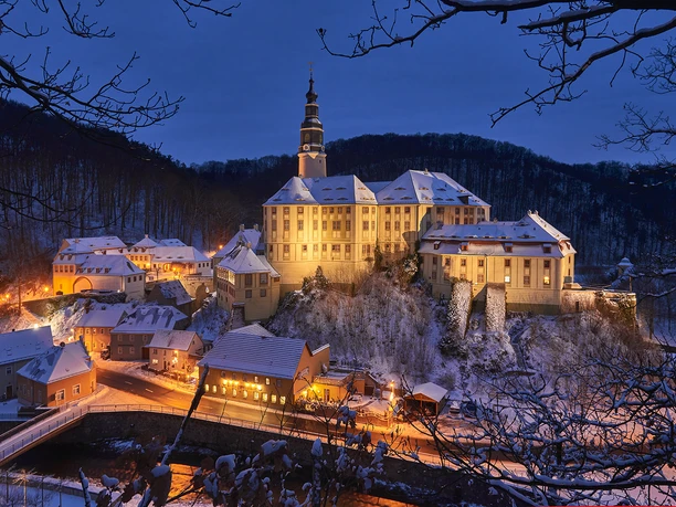 Schloss Weesenstein im Winter Beleuchtetes Schloss Weesenstein auf einem verschneiten Hügel bei Dämmerung, umgeben von kleinen Häusern und einem Fluss, winterliche Atmosphäre.