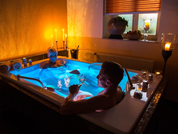 Two people relax by candlelight in a blue whirlpool bathtub, holding glasses of champagne, surrounded by towels and candles.