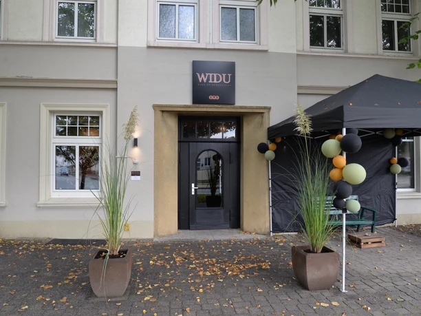 Außenansicht eines Gebäudes mit einem Schild "WIDU" über der Eingangstür, dekoriert mit Pflanzen.