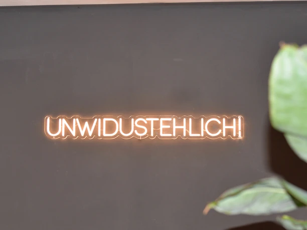 Leuchtende Neonaufschrift "UNWIDUSTEHLICH" auf dunklem Hintergrund, rechte Bildseite mit Pflanze.