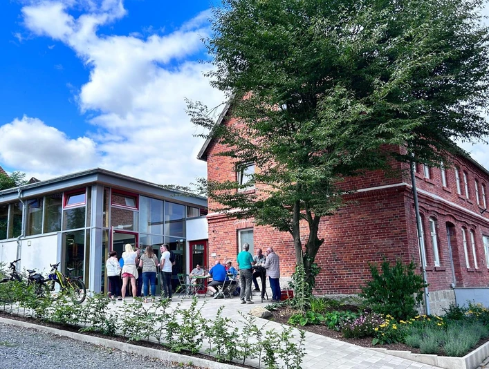 Aus Krus Bellersen - Außenansicht Das Bild zeigt ein renoviertes Backsteingebäude mit moderner Glasfassade und versammelten Personen.