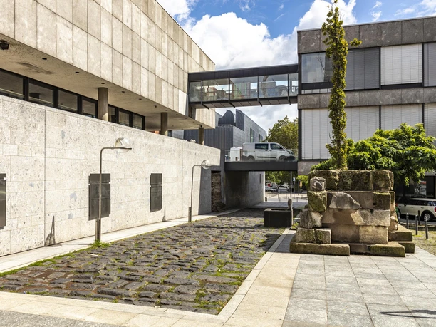 Römische Hafenstraße Blick auf eine moderne Straßenbrücke und Mauer aus hellem Stein, umgeben von historischen Pflastersteinen und einem antiken römischen Steinsockel