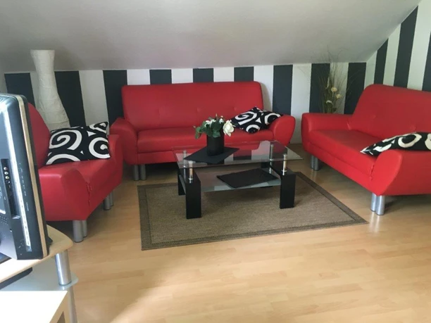 Wohnzimmer FeWo Brüggemann Wohnzimmer mit drei roten Sofas, Glastisch, dekorativen Kissen, moderner Stil, helle Holzböden.