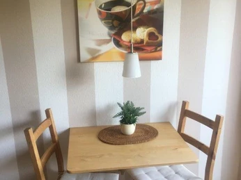 Esszimmer FeWo Brüggemann Kleiner Essbereich mit Holztisch, zwei gepolsterten Stühlen, einem Pflanzenarrangement und einem Wandbild.