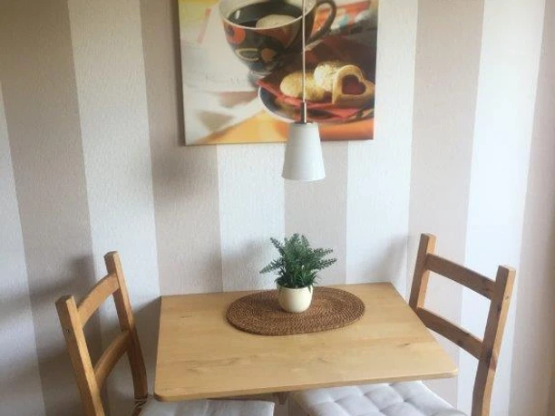Esszimmer FeWo Brüggemann Kleiner Essbereich mit Holztisch, zwei gepolsterten Stühlen, einem Pflanzenarrangement und einem Wandbild.