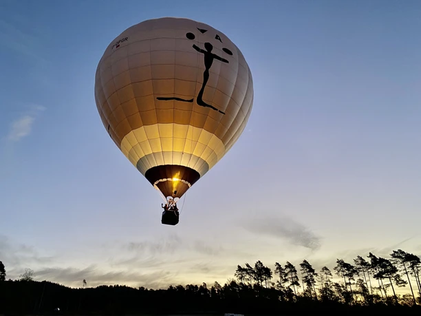 Die Welt von oben erleben! Heißluftballon mit Menschengrafik erhebt sich bei Sonnenuntergang über eine bewaldete Landschaft.