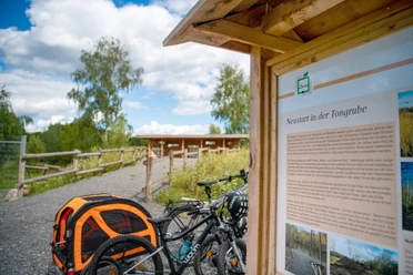 Neustart in der Tongrube.jpg Fahrräder und ein Hundewagen vor einer Info-Tafel in einer landschaftlichen Naturanlage.