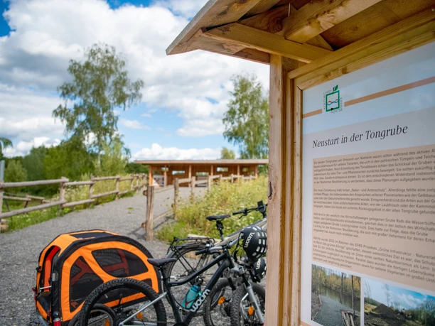 Neustart in der Tongrube.jpg Fahrräder und ein Hundewagen vor einer Info-Tafel in einer landschaftlichen Naturanlage.