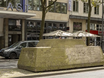 Motionless traffic Großer, grauer Betonsockel am Straßenrand in Köln, flankiert von parkenden Autos und Geschäften.Large, gray concrete pedestal on the side of the road in Cologne, flanked by parked cars and stores.