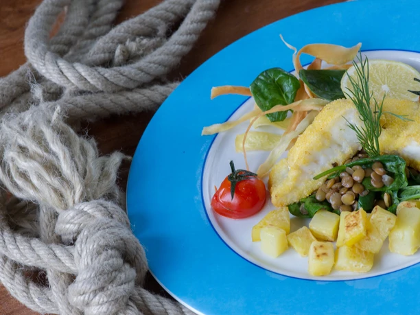 A plate with breaded fish, diced potatoes, lentils, spinach, tomatoes and a slice of lemon on a blue rim, next to a rope on a wooden table.