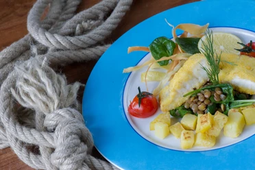 Bio-Restaurant StrandGut Un piatto con pesce impanato, cubetti di patate, lenticchie, spinaci, pomodori e una fetta di limone su un bordo blu, accanto a una corda su un tavolo di legno.