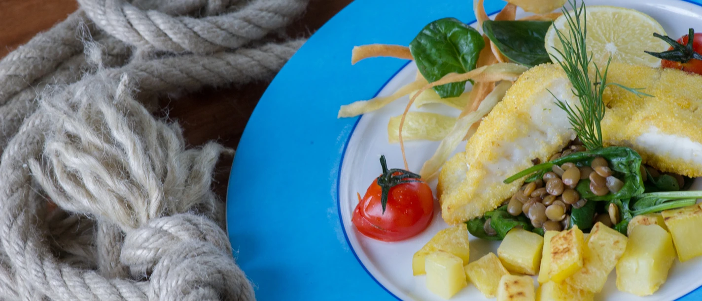 Bio-Restaurant StrandGut Talerz z panierowaną rybą, kostkami ziemniaków, soczewicą, szpinakiem, pomidorami i plasterkiem cytryny na niebieskiej obręczy, obok liny na drewnianym stole.