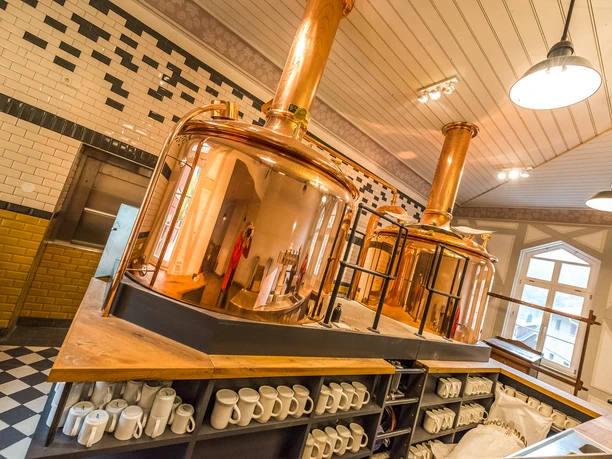 Braukessel Brau-Manufaktur Kupferne Braukessel in einer Brauerei mit weißen und schwarzen Fliesen an den Wänden; darunter Regale mit weißen Krügen.