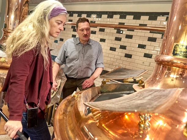 Twee mensen kijken naar een grote koperen brouwinstallatie in een brouwerij met betegelde muren; de sfeer is geconcentreerd.