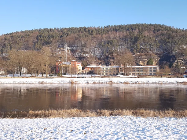 Sendigs Wintergarten im Parkhotel Flussufer im Winter mit schneebedecktem Boden, dahinter bunte Gebäude und ein bewaldeter Hügel unter klarem, blauem Himmel.