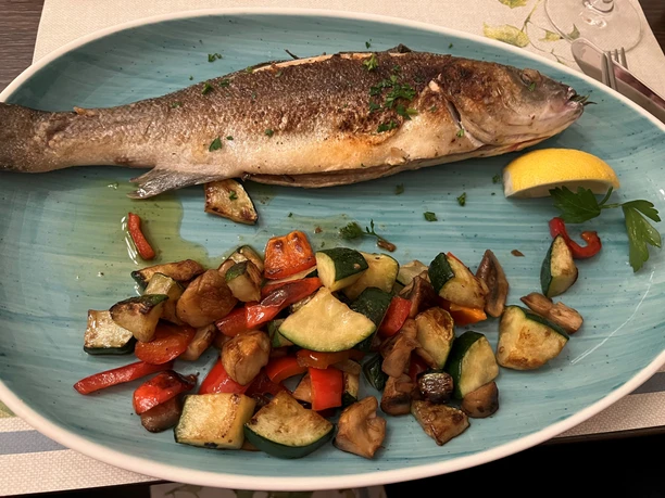 Grilled fish with crispy skin on a blue plate, accompanied by roasted vegetables and lemon wedge.