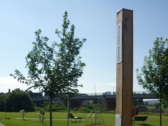 Ein Holzpfahl mit Bibelspruch in einem Park, umgeben von Bäumen und einer Brücke im Hintergrund.