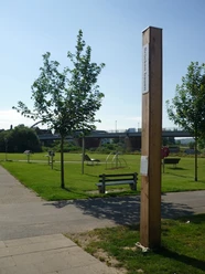 Bibel-Pfahl in Beverungen Ein Holzpfahl mit Bibelspruch in einem Park, umgeben von Bäumen und einer Brücke im Hintergrund.