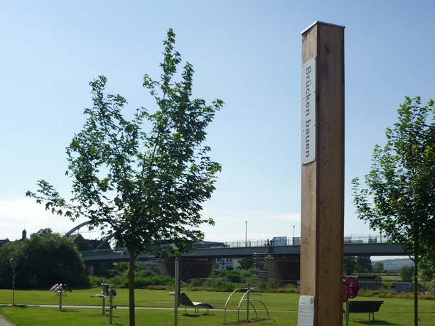Bibel-Pfahl in Beverungen Ein Holzpfahl mit Bibelspruch in einem Park, umgeben von Bäumen und einer Brücke im Hintergrund.