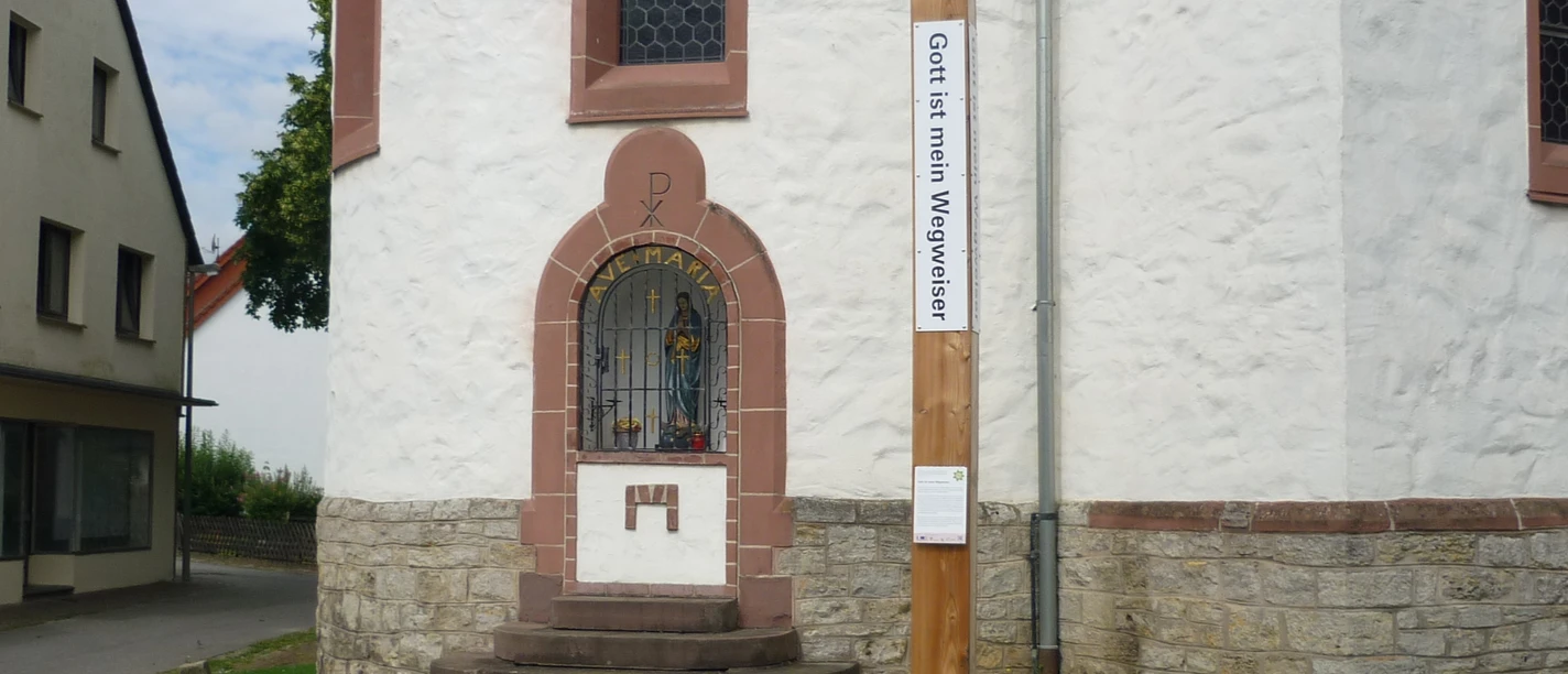 Runder Kirchturm mit steinernen Stufen, bunt verglastes Fenster, daneben ein hölzerner Bibel-Pfahl.