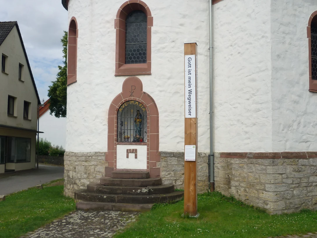 Bibel-Pfahl in Hembsen Runder Kirchturm mit steinernen Stufen, bunt verglastes Fenster, daneben ein hölzerner Bibel-Pfahl.