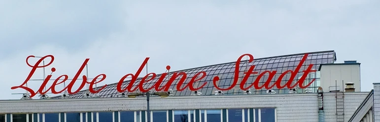 LiebedeineStadt Gebäude mit rotem Schriftzug „Liebe deine Stadt“ auf dem Dach, bewölkter Himmel im Hintergrund.