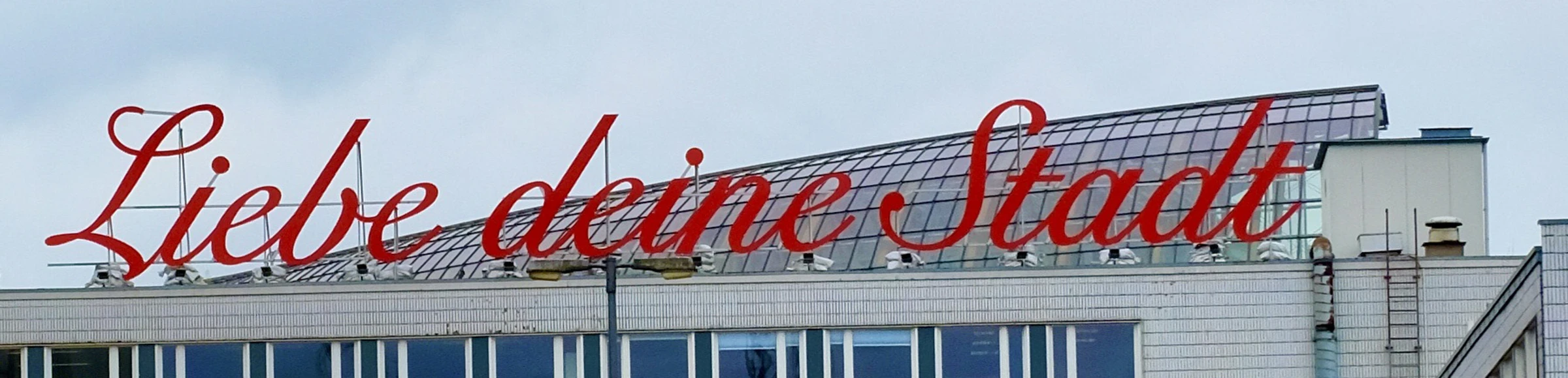 LiebedeineStadt Gebäude mit rotem Schriftzug „Liebe deine Stadt“ auf dem Dach, bewölkter Himmel im Hintergrund.Building with red lettering “'Liebe deine Stadt”' on the roof, cloudy sky in the background.