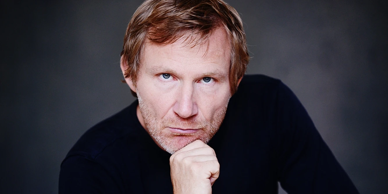 Porträt eines nachdenklichen Mannes mittleren Alters mit verschränkten Fingern und ernstem Blick.