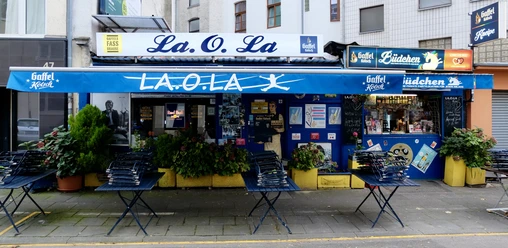 La Ola Büdchen Kiosk "La.O.La" in Köln, mit blauer Fassade und Gaffel-Logo markanten Sitzmöglichkeiten vor dem Eingang.