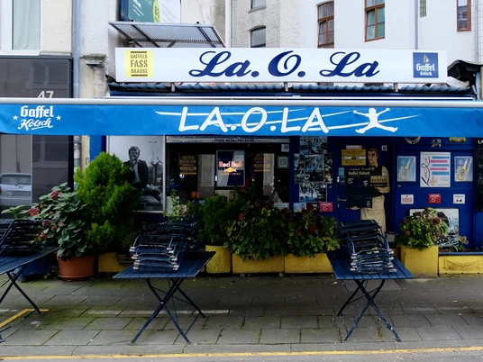 La Ola Büdchen Kiosk "La.O.La" in Köln, mit blauer Fassade und Gaffel-Logo markanten Sitzmöglichkeiten vor dem Eingang.Kiosk "La.O.La" in Cologne, with blue façade and Gaffel logo, striking seating in front of the entrance.