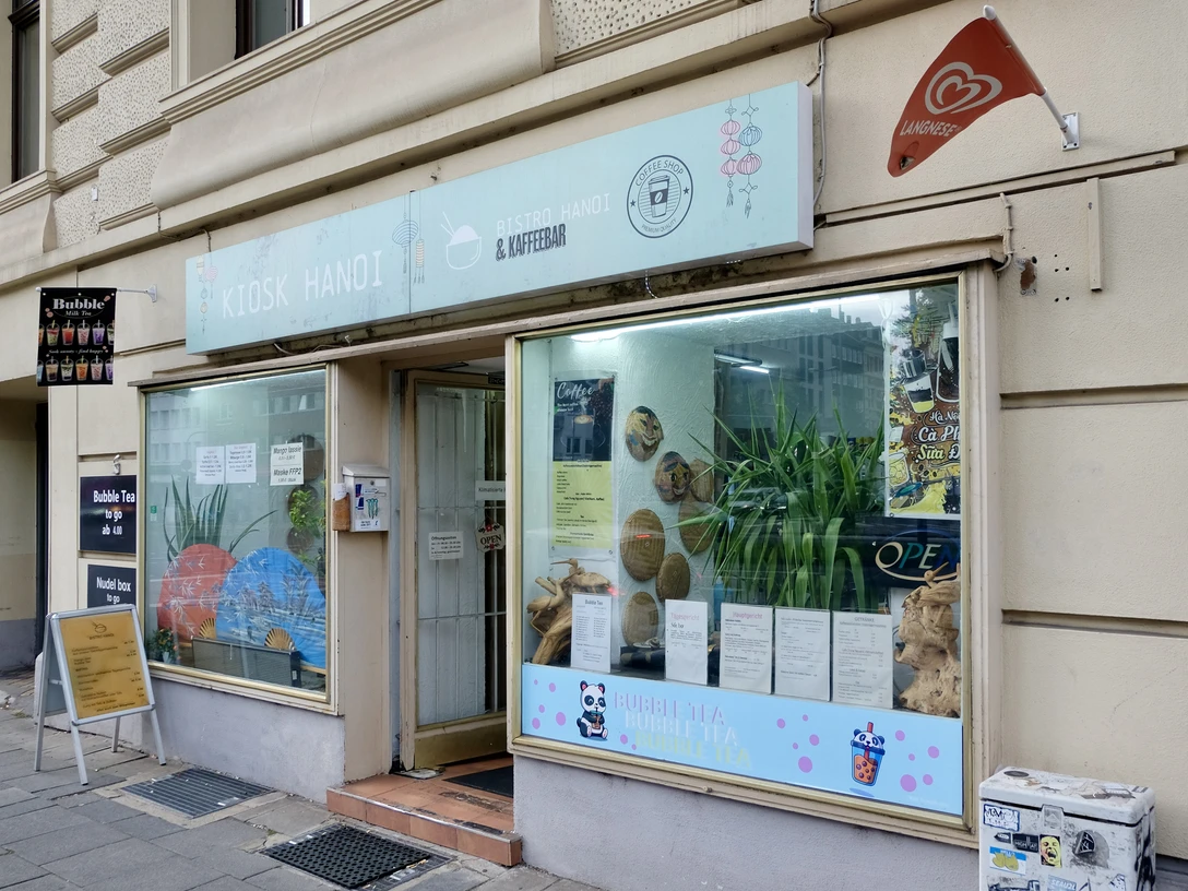 Kiosk Hanoi Die Fassade ist mit großen Fenstern ausgestattet, hinter denen Dekorationen und Pflanzen zu sehen sind. Ein Schild mit dem Namen des Geschäfts und Informationen über das angebotene Sortiment befindet sich oberhalb der Fenster. Vor dem Kiosk steht ein Aufsteller mit weiteren Details zu den angebotenen Produkten. Die Fenster sind mit Illustrationen von Bubble Tea verziert.The façade has large windows, behind which decorations and plants can be seen. A sign with the name of the store and information about the range on offer is located above the windows. In front of the kiosk is a display with further details about the products on offer. The windows are decorated with illustrations of bubble tea.