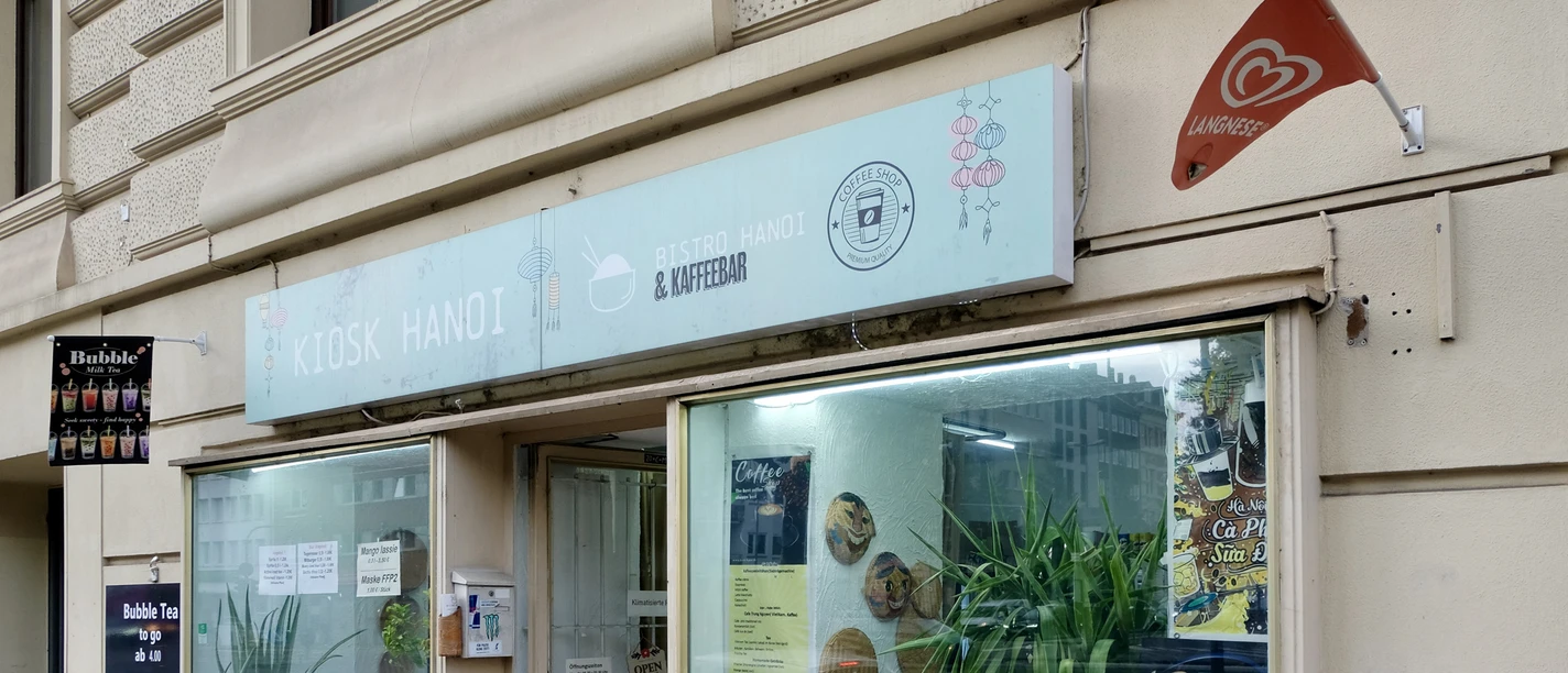 Kiosk Hanoi The façade has large windows, behind which decorations and plants can be seen. A sign with the name of the store and information about the range on offer is located above the windows. In front of the kiosk is a display with further details about the products on offer. The windows are decorated with illustrations of bubble tea.