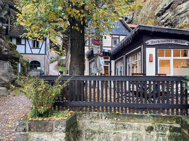 Hrázděný dům s nápisem "Rathewalder Mühle", obklopený podzimními stromy a skalami, s cestou v popředí.