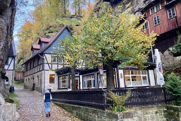 Vakwerkhuis in een herfstlandschap, omringd door bomen met gele bladeren; een persoon loopt over een pad.