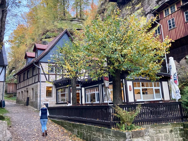 Hrázděný dům v podzimní krajině, obklopený stromy se žlutým listím; po cestě kráčí člověk.