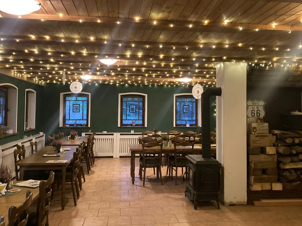 Rathewalder Mühle Kaminstube Gemütlicher Raum mit Holztischen und -stühlen, Lichterketten an der Decke, grüne Wände, Kaminofen und gestapeltes Holz; Route 66 Schild an der Wand.