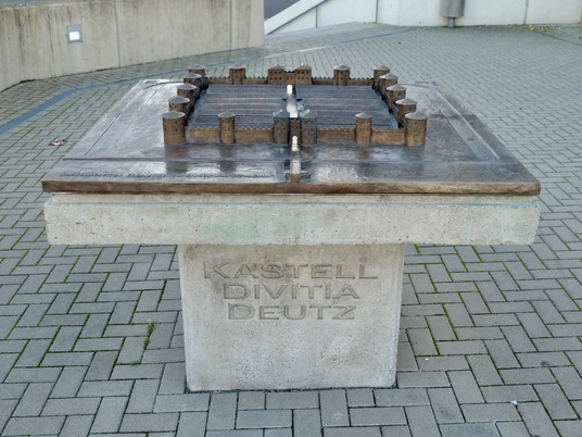 Divitia-Deutz fort Das Bild zeigt ein modernes Modell des antiken Kastells Divitia in Köln-Deutz. Es ist aus Metall gefertigt und hebt sich kontrastreich von der hellen Steinbasis ab. Das Modell liegt auf einem quadratischen Sockel, der mit der Inschrift "KASTELL DIVITIA DEUTZ" versehen ist. The picture shows a modern model of the ancient Divitia fort in Cologne-Deutz. It is made of metal and contrasts sharply with the light-colored stone base. The model rests on a square base with the inscription "KASTELL DIVITIA DEUTZ".