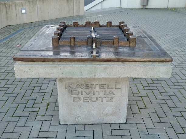 Kastell Divitia-Deutz Das Bild zeigt ein modernes Modell des antiken Kastells Divitia in Köln-Deutz. Es ist aus Metall gefertigt und hebt sich kontrastreich von der hellen Steinbasis ab. Das Modell liegt auf einem quadratischen Sockel, der mit der Inschrift "KASTELL DIVITIA DEUTZ" versehen ist.