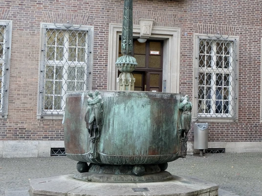 Carnival fountain Der Fastnachtsbrunnen in Köln zeigt eine künstlerische Skulptur mit Bronzefiguren, die die berühmte fünfte Jahreszeit darstellt. Der Brunnen ist ein Kunstwerk mit detaillierten Figuren, das auf einem gepflasterten Vorplatz vor einem Backsteingebäude steht. Eine Säule in der Mitte, gekrönt mit symbolischen Karnevalsfiguren, zieht den Blick auf sich. Umrahmt von sechs Bronzemasken, steht es stellvertretend für Fröhlichkeit und Tradition des rheinischen Karnevals.The Fastnachtsbrunnen in Cologne features an artistic sculpture with bronze figures depicting the famous fifth season. The fountain is a work of art with detailed figures that stands on a paved forecourt in front of a brick building. A column in the middle, crowned with symbolic carnival figures, draws the eye. Framed by six bronze masks, it represents the cheerfulness and tradition of the Rhineland carnival.
