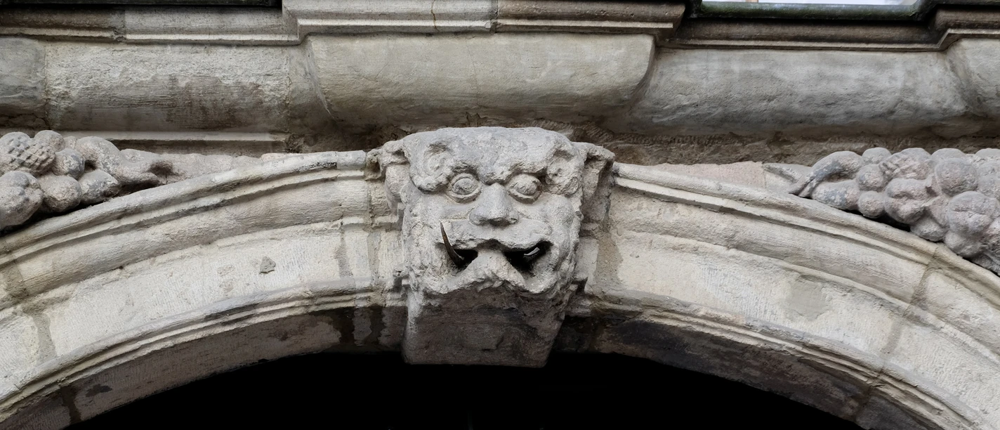 Grinkopf Stone grin head with artistic grimace above an arch, framed by fruit motifs.