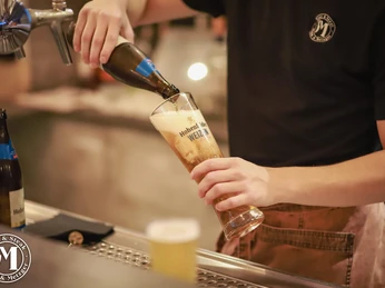 Mann und Metzger Bier Ein Kellner füllt ein Hefeweizen in ein hohes Glas an einer Bar. Die Marke "Hofbräu München" ist sichtbar.
