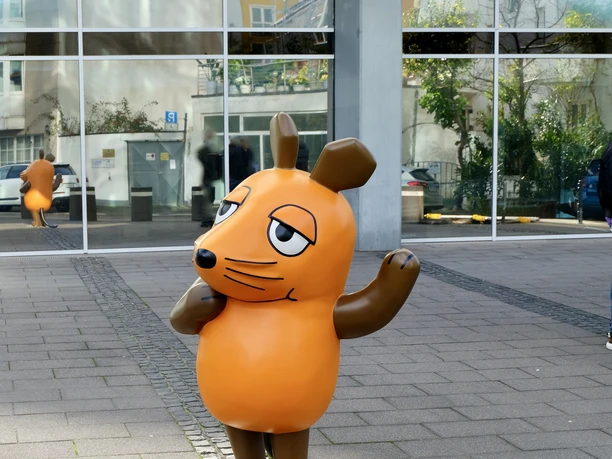 Mouse Statue in Cologne Orange mouse statue with a raised paw in front of a reflective glass facade in Cologne