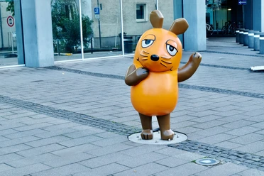 Maus Statue Die Maus-Statue mit erhobenem Arm, ein Symbol der beliebten Kindersendung, steht auf Gehwegplatten vor einem modernen Gebäude in Köln.