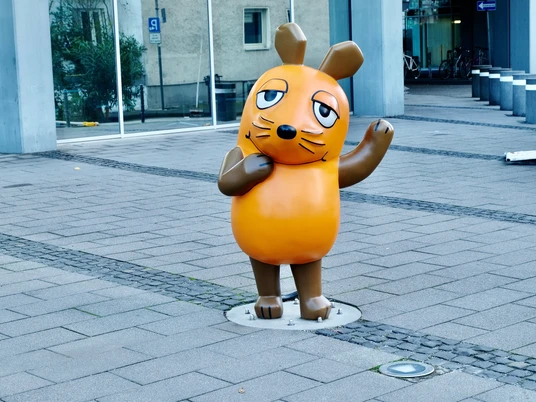 Mouse statue Die Maus-Statue mit erhobenem Arm, ein Symbol der beliebten Kindersendung, steht auf Gehwegplatten vor einem modernen Gebäude in Köln.The mouse statue with raised arm, a symbol of the popular children's show, stands on paving slabs in front of a modern building in Cologne.