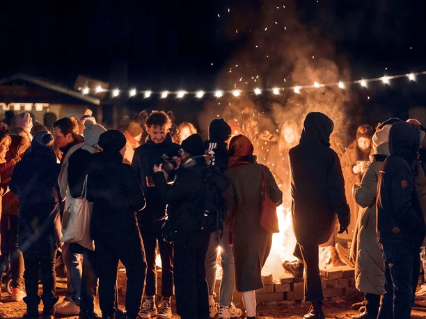 Veihnachtsmarkt-RosaRot-eV_2.jpg Menschen stehen abends an einem offenen Feuer unter Lichterketten und wärmen sich gemeinsam