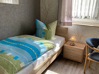 FeWo Schniedermann_Beispielzimmer 1_CC-BY-SA_Teutoburger_Wald_Stadt_Schloss_Holte-Stukenbrock.jpg Ein Einzelbett in einem hellen Zimmer mit Holzboden, modernem Dekor und einem Nachttisch mit Lampe.